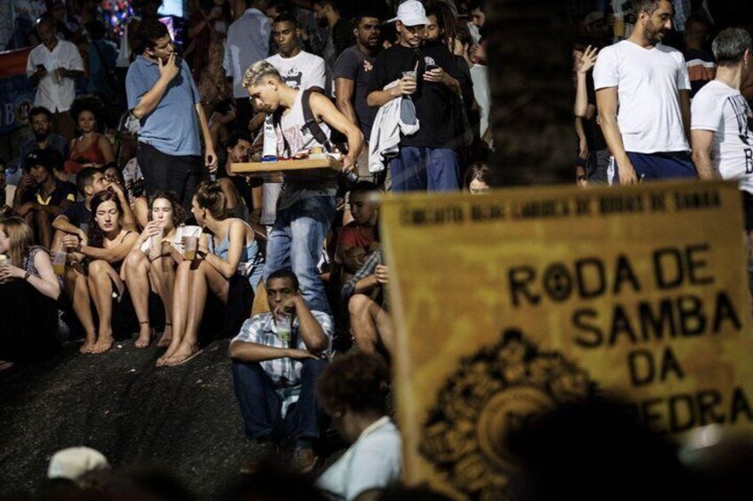 A crowd of people, some sitting and some standing, with a sign that reads 'RODA DE SAMBA DA REDRA'. 2