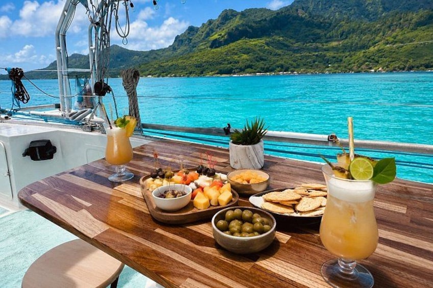 A wooden table with food and drinks on a boat with a turquoise sea and green hills in the background. 5