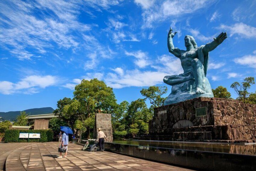 A large bronze statue of a figure with outstretched arms, a stone pedestal, and a paved walkway with a person holding an umbrella. 5