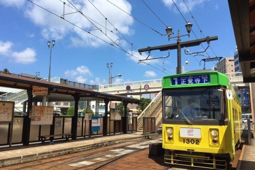 A yellow tram at a station with overhead wires, a pedestrian bridge, and a clear sky. 1