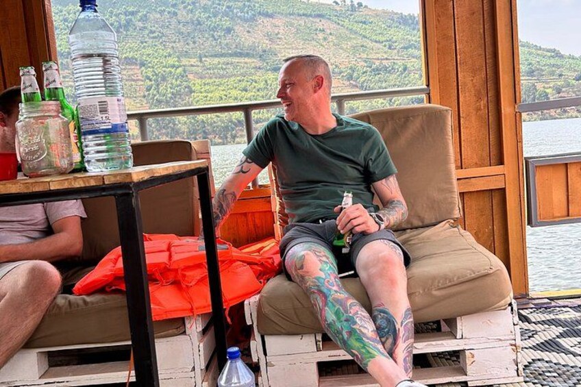 A man with tattoos sitting on a wooden bench by a window overlooking a body of water. 2