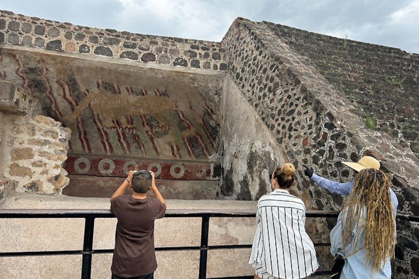 A stone wall with ancient frescoes, a person taking a photo, and another pointing at the wall. 4