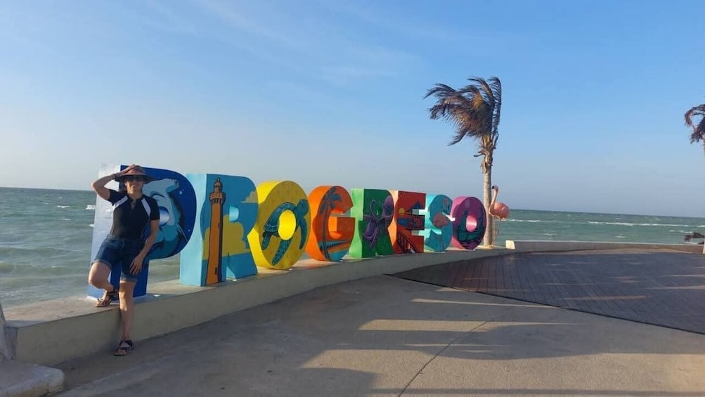 A person standing in front of a colorful sign with the word 'PROGRESO' and palm trees in the background. 3