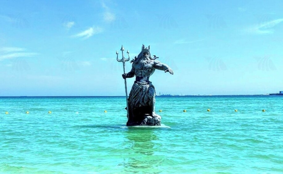 A statue of a mythological figure standing in the water with a trident, surrounded by clear blue sea and distant shoreline. 4
