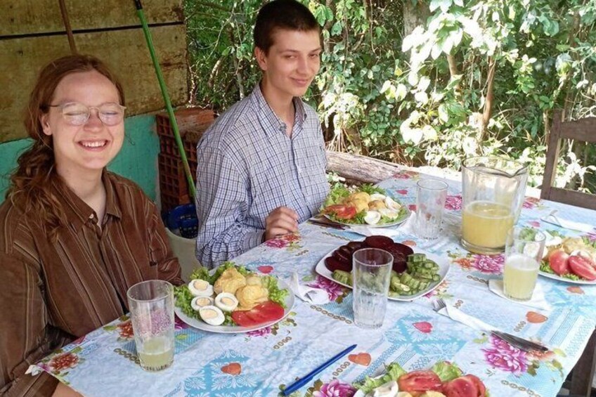 Two people sitting at an outdoor table with a floral tablecloth, plates of food, and drinks. 3