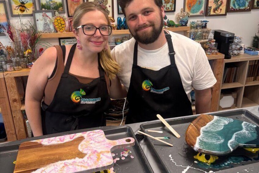 Two people wearing aprons with a logo, standing in front of a workbench with a wooden board and a tray with a blue liquid. 1