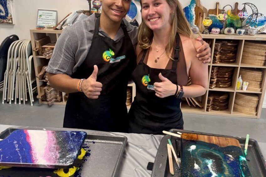 Two people wearing aprons with a logo, standing in a workshop with art supplies and pottery on shelves. 2