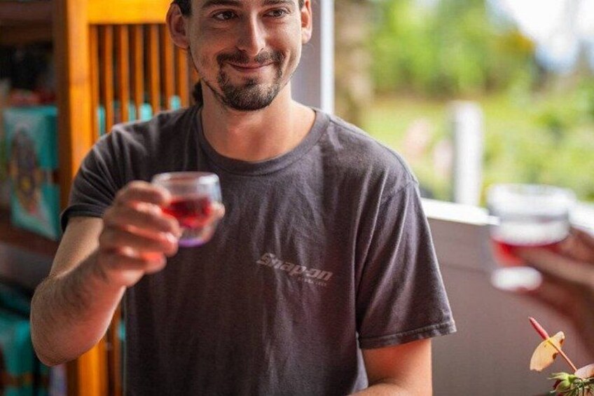A person holding a glass of red liquid, wearing a dark t-shirt with a logo, in an indoor setting with a blurred background. 1