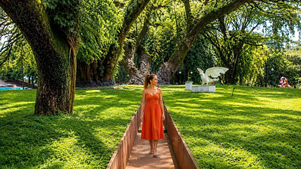 A woman in an orange dress walking on a wooden bridge in a park with green grass, trees, and sculptures. 3