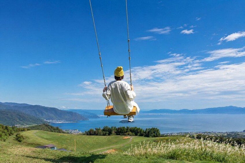 A person on a swing overlooking a coastal landscape with hills and a view of the sea. 1