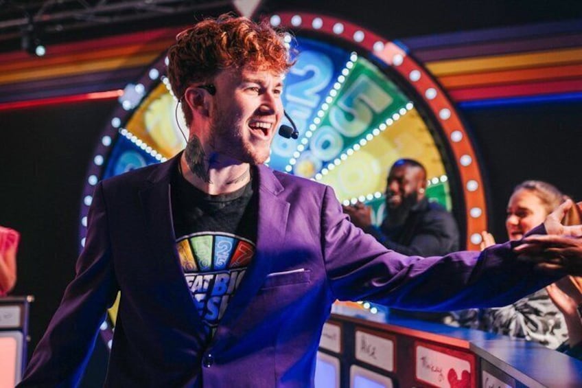 A person in a purple suit with a microphone, standing in front of a colorful, circular background with lights. 2