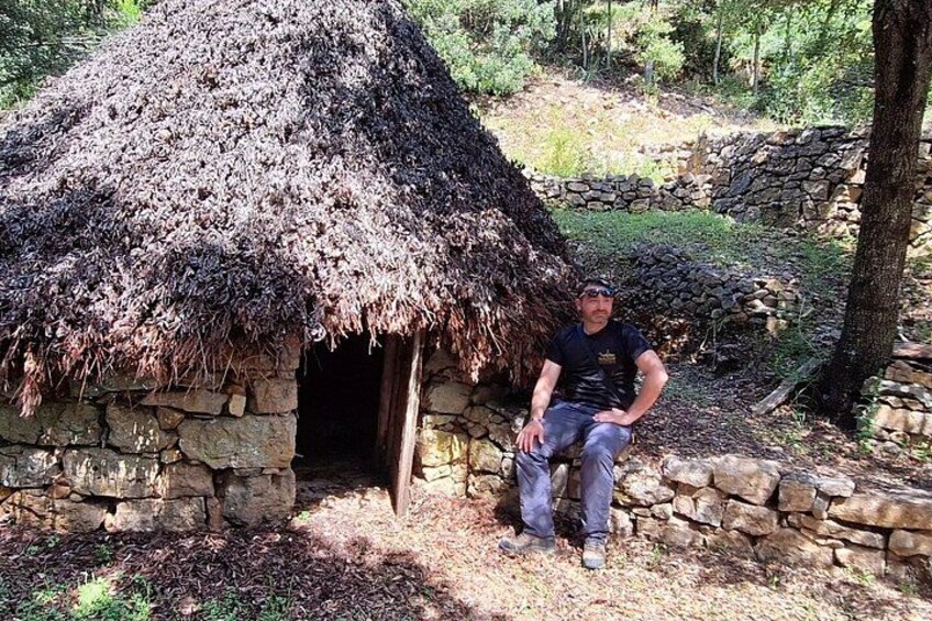 A man sitting outside a thatched-roof hut with stone walls, surrounded by trees and a stone wall in the background. 1