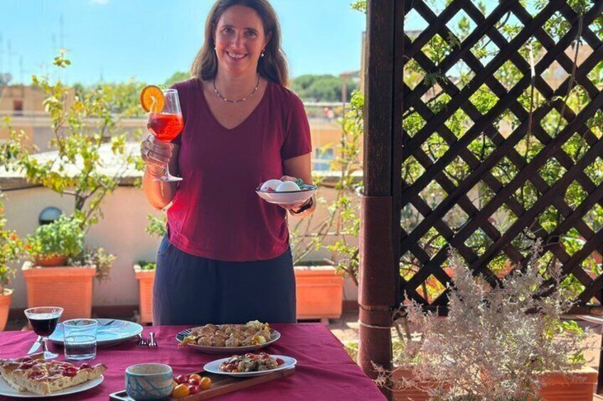 A person holding a glass of red drink and a plate with food, standing at a table with dishes and a lattice structure in the background. 4