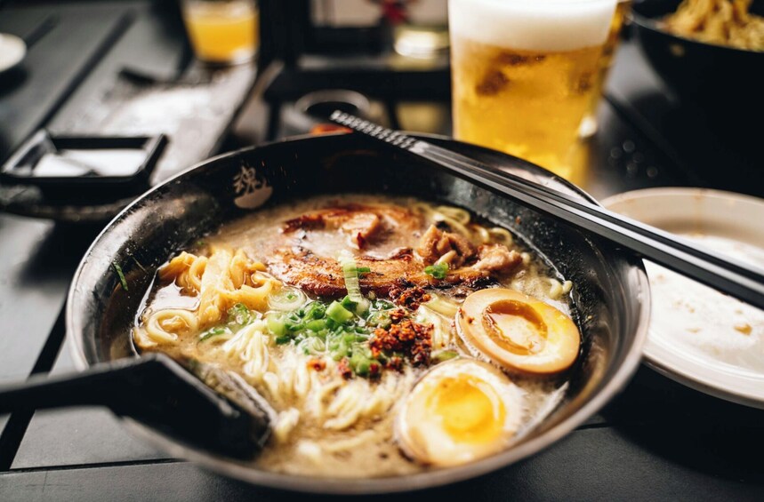 A bowl of ramen with noodles, meat, and toppings, accompanied by a glass of beer and chopsticks on a table. 1