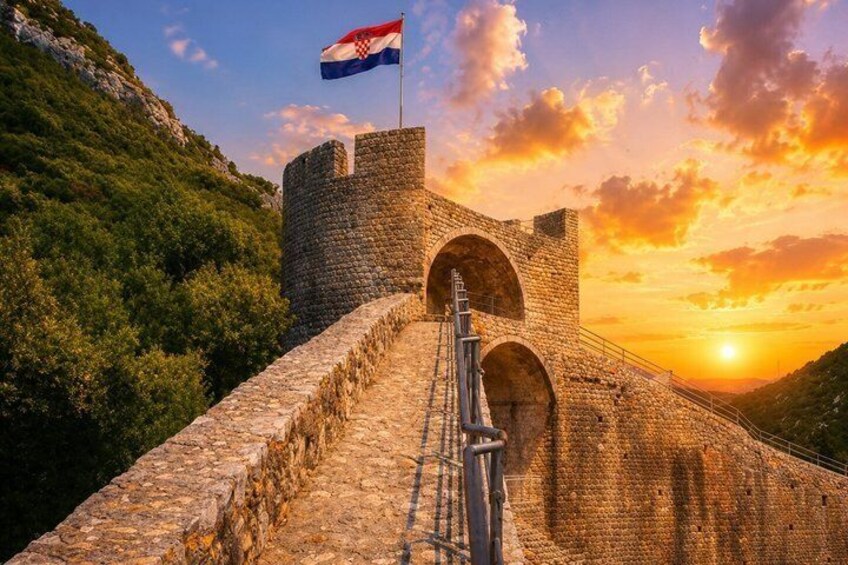 Un muro de piedra con aberturas en arco, un asta con una bandera, y una puesta de sol al fondo. 4
