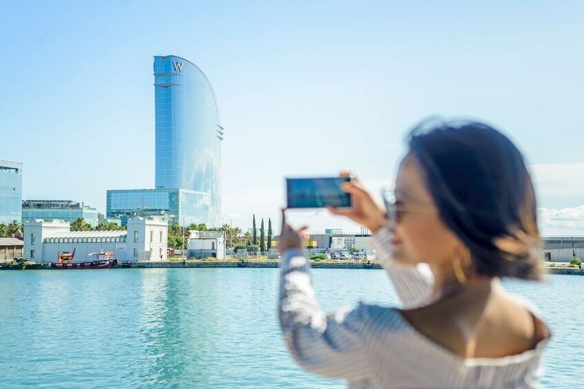 A person taking a photo with a smartphone, a modern skyscraper, and a calm body of water. 2