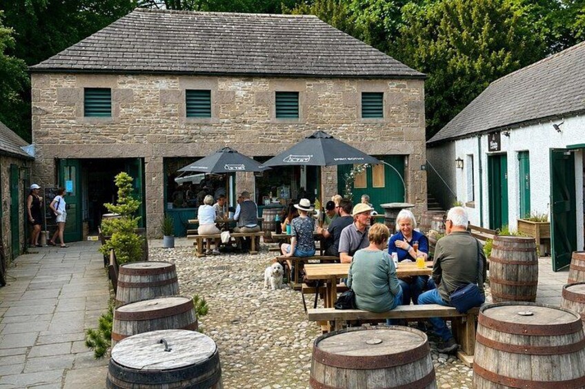 An outdoor café with stone buildings, wooden barrels, and people dining. 2