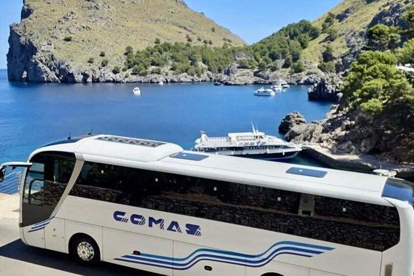 Een witte bus met blauwe strepen geparkeerd nabij water met boten en rotsachtig terrein op de achtergrond. 2