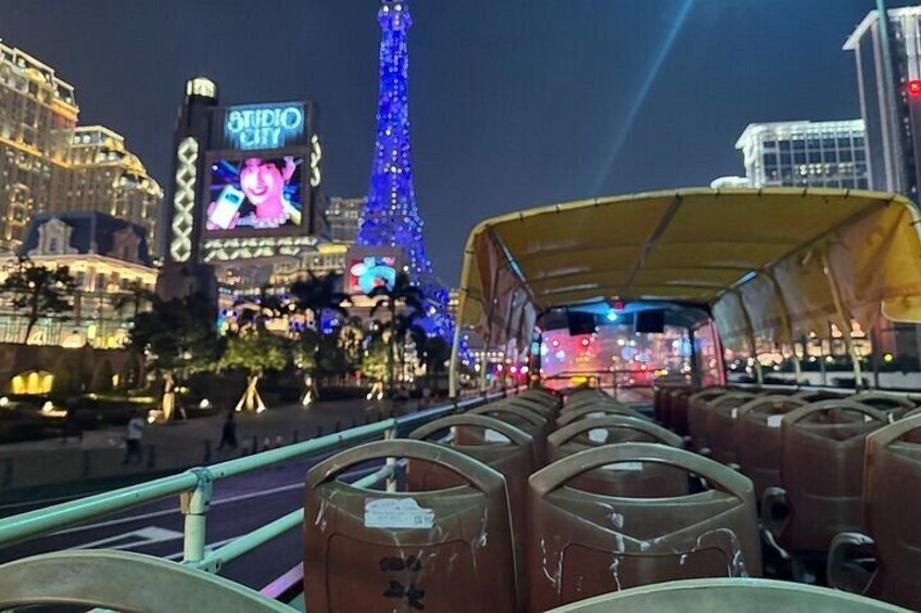 A nighttime cityscape with illuminated buildings, a large digital billboard, and a covered bus in the foreground. 1