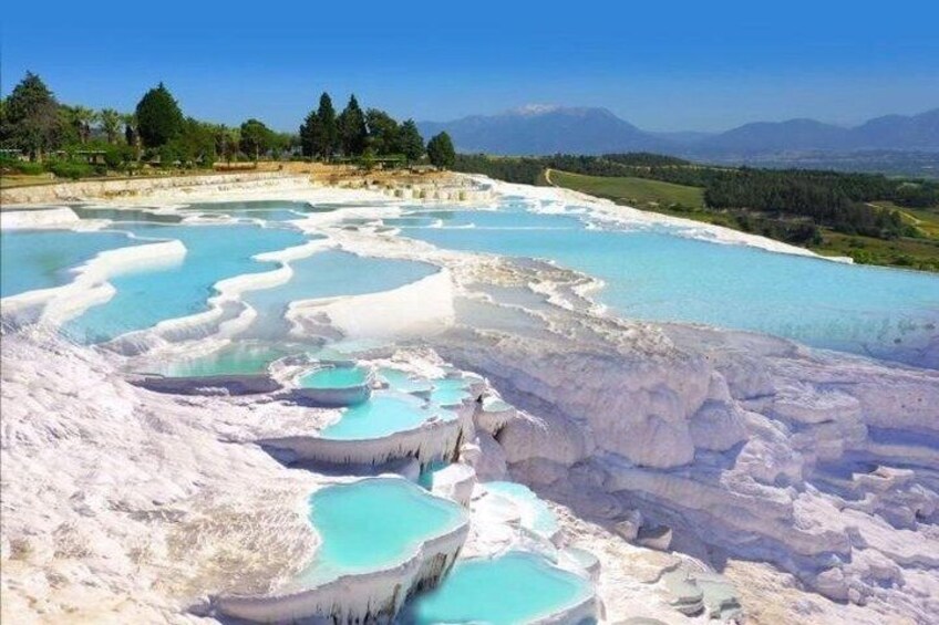 A landscape with terraced pools of turquoise water, white mineral deposits, and a distant mountain range. 1