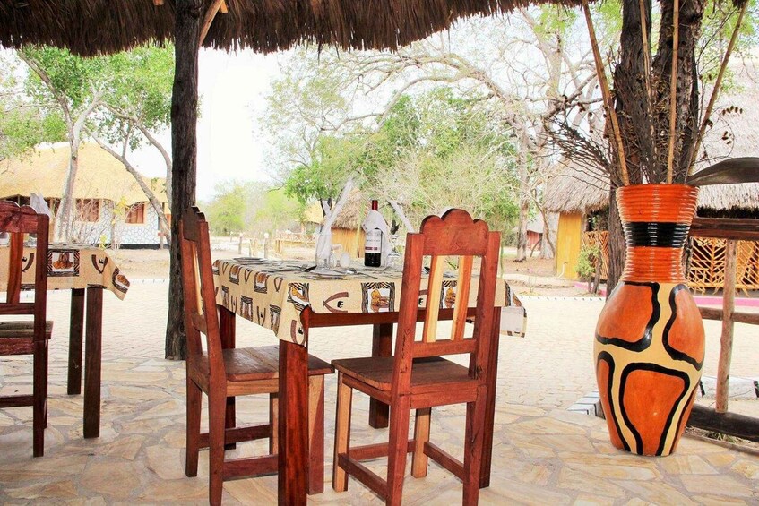 An outdoor dining area with wooden chairs and tables, a large decorative vase, and thatched-roof huts in the background. 5