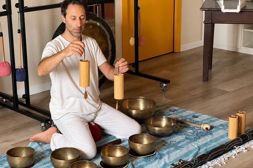 A person sitting on a mat surrounded by several brass bowls and wooden instruments, likely used for meditation or sound therapy. 5