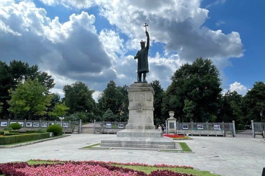 A statue of a figure holding a cross on a pedestal, surrounded by a paved area with flower beds and a fence. 5
