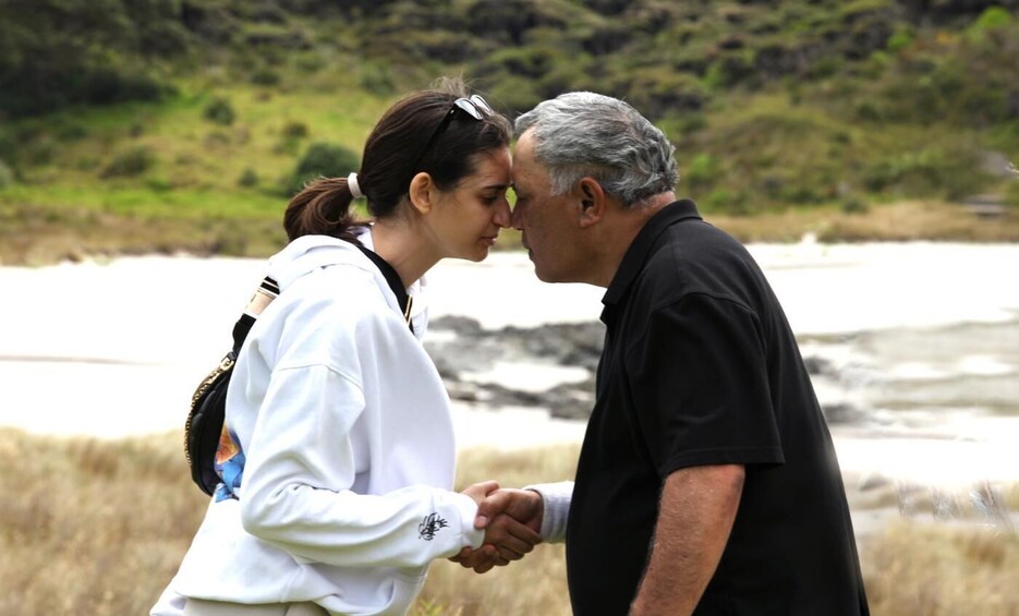 Two people shaking hands in a natural outdoor setting with a body of water and grassy hills in the background. 3