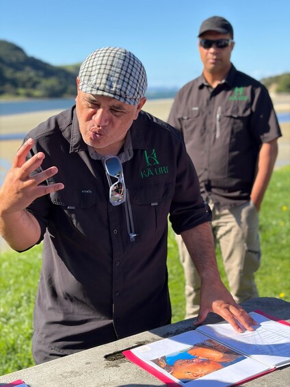 A person gesturing at a table with papers and a book, outdoors with a body of water and hills in the background. 2
