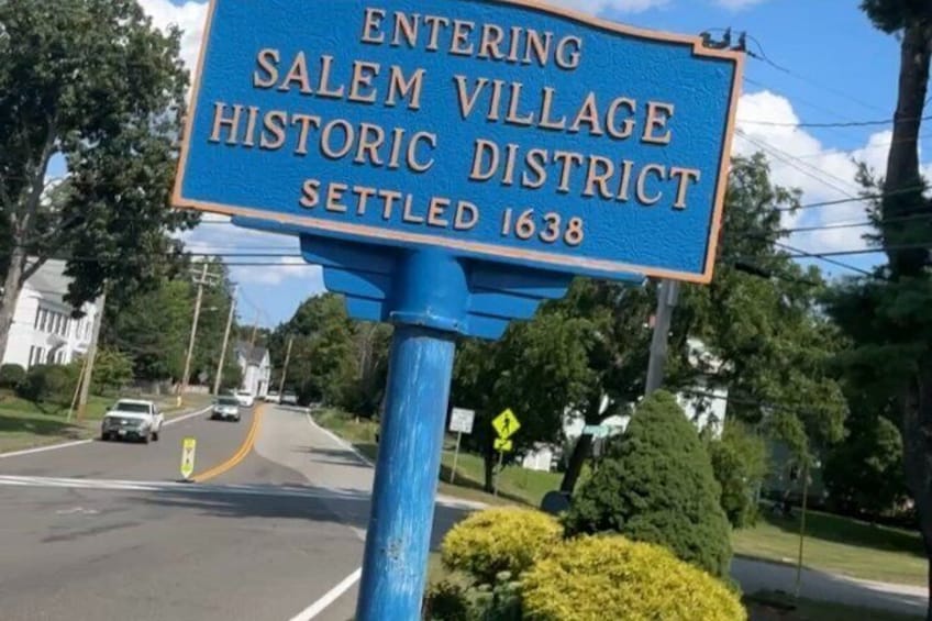 A blue sign with white text indicating the entrance to Salem Village Historic District, settled in 1638. 5