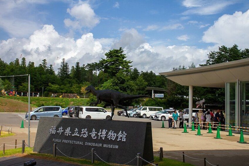 A dinosaur museum entrance with a large dinosaur statue, cars, and people. 4