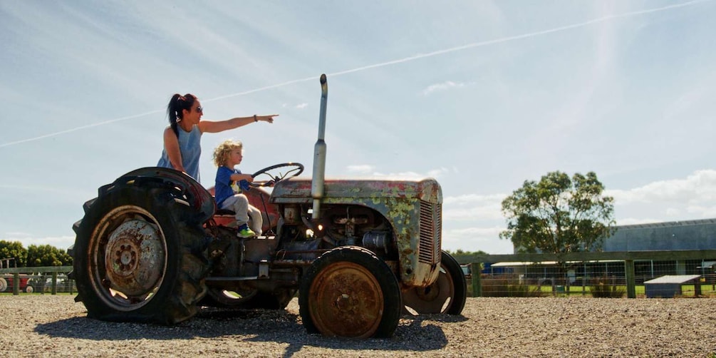 A vintage tractor with a child driving, a woman pointing, and a rural landscape with a fence and trees in the background. 5