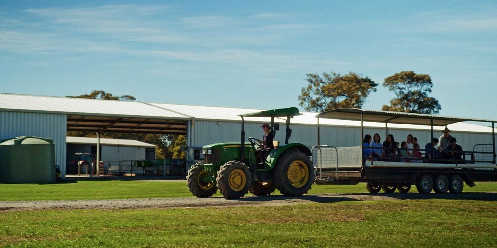 A green tractor pulling a trailer with people seated on it, parked in front of a large industrial building with a metal roof. 1