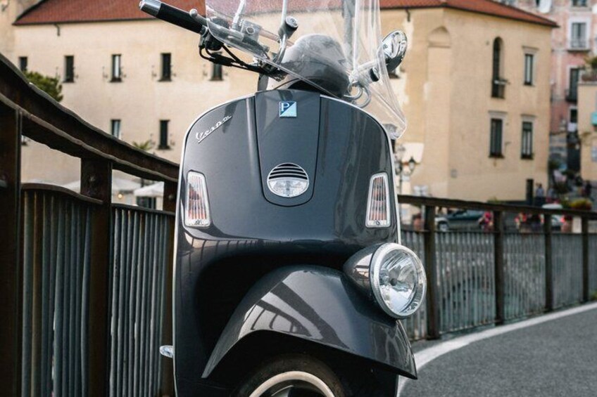 A black Vespa scooter parked on a street with a metal fence and buildings in the background. 4