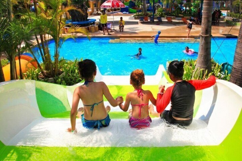 A water slide with three people at the bottom, a pool with people in the background, and palm trees around. 3
