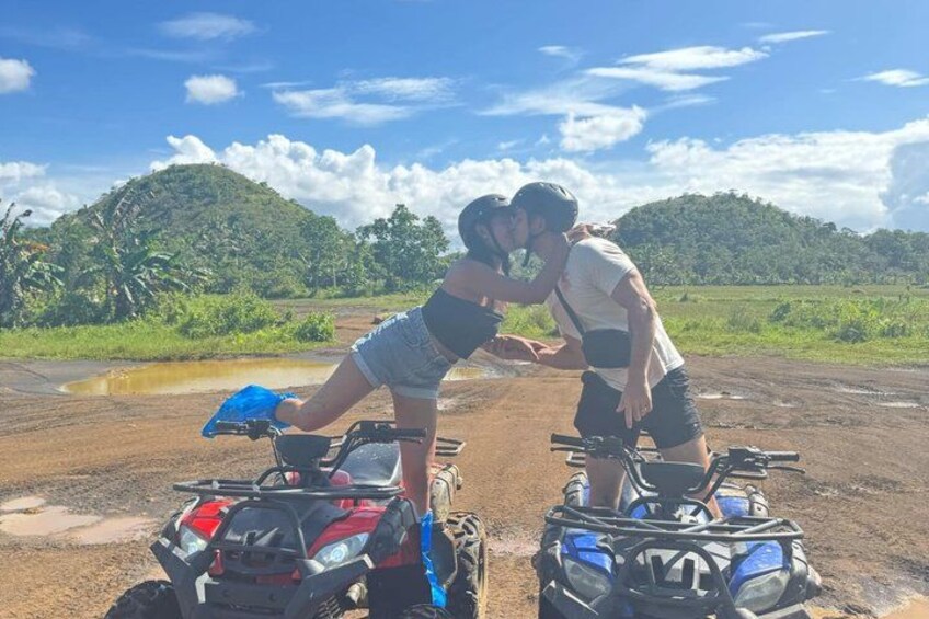 Two people on ATVs in a rural setting with a dirt road and greenery in the background. 4