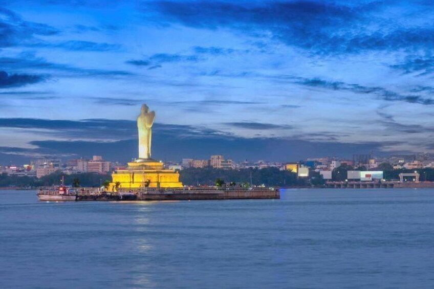 A large illuminated statue on a platform in the water, with a cityscape in the background. 1