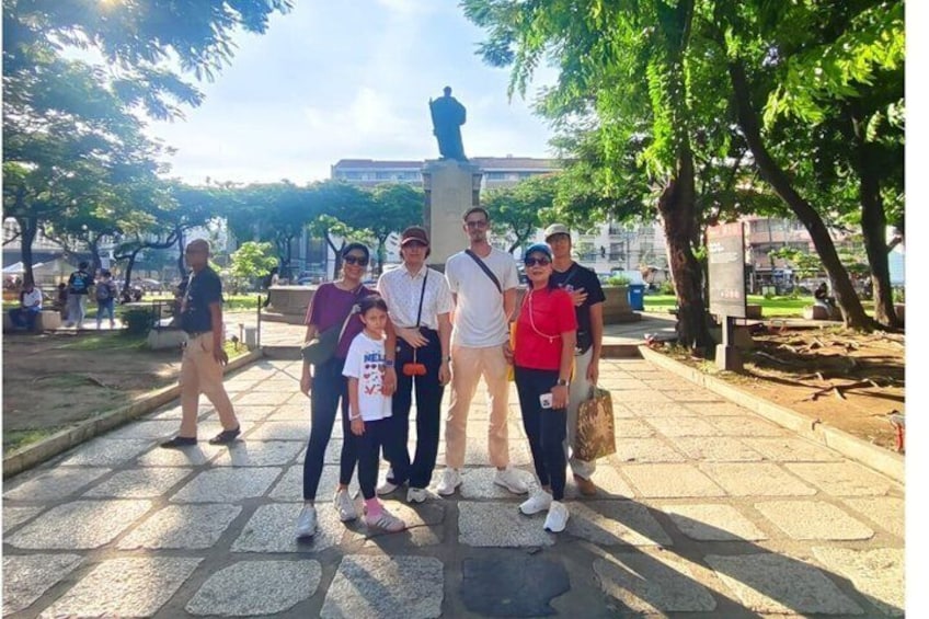 A group of people posing in front of a statue in a park with trees and a paved walkway. 1