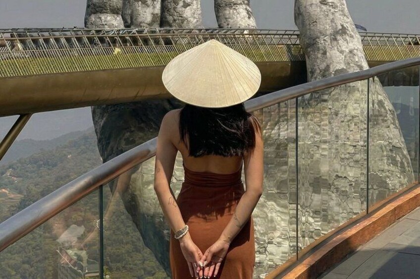 A person wearing a conical hat standing on a glass bridge with a railing, overlooking a mountainous landscape. 1