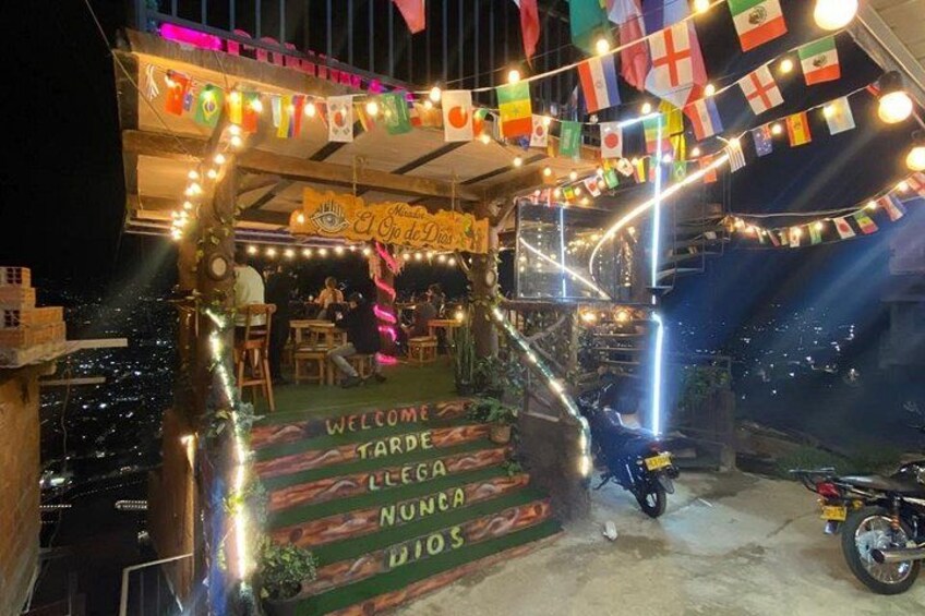 A brightly lit outdoor seating area with flags, a staircase, and a sign that reads 'WELCOME TARDES NUNCA DIOS.' 1