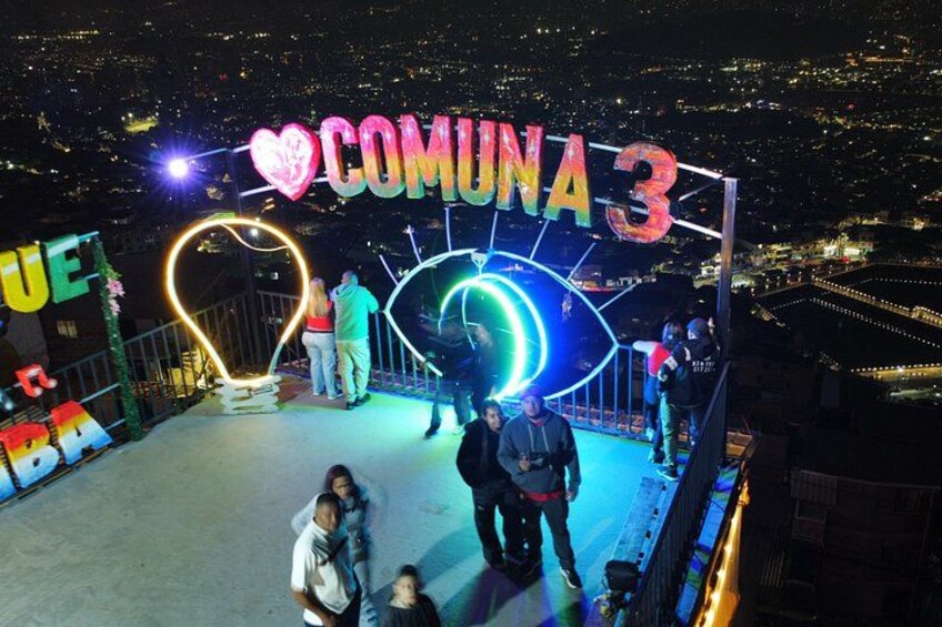 A nighttime view of a cityscape with a large illuminated sign reading 'COMUNA 3' and a heart-shaped light installation. 5