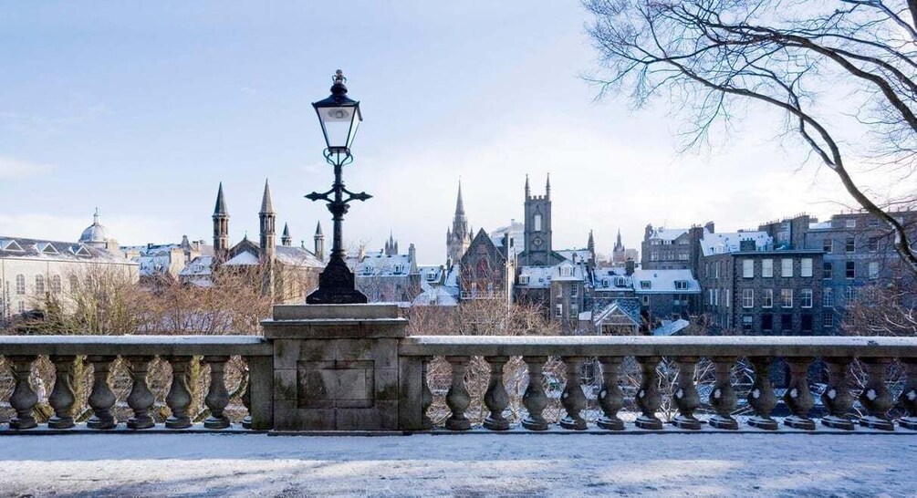 A snow-covered bridge with a stone balustrade and a street lamp, overlooking a cityscape with historic buildings and spires. 1