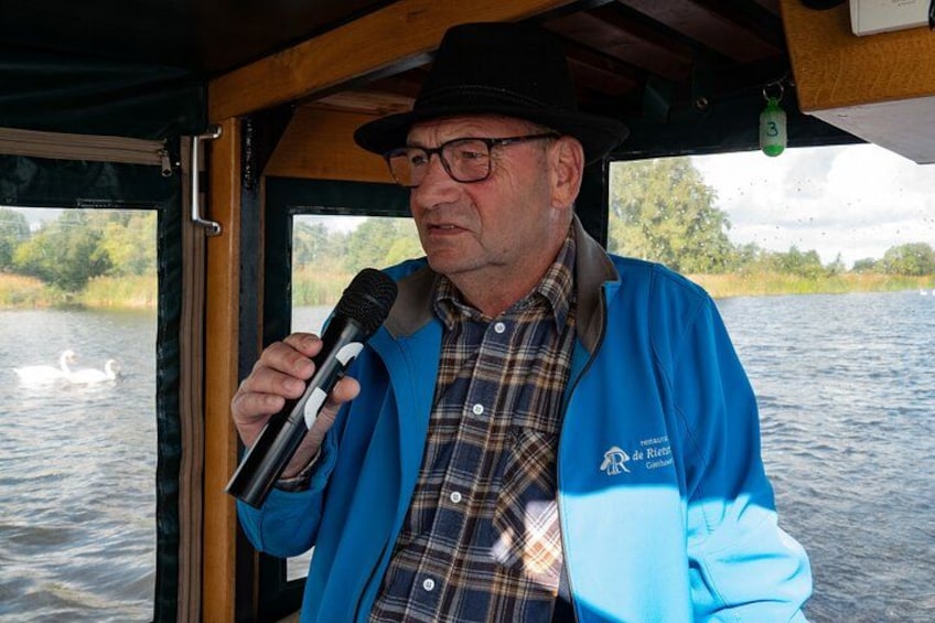 Una persona sosteniendo un micrófono, vestida con una chaqueta azul con un logotipo, de pie en un barco, con un cuerpo de agua y follaje en el fondo. 5