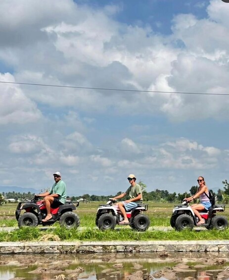 Three people riding ATVs on a rural road with fields and a water body on the side. 3
