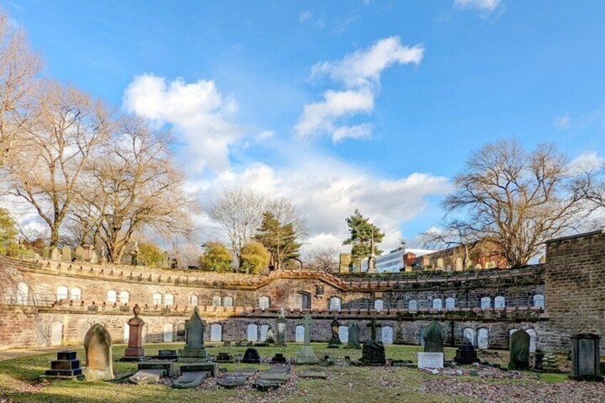 A cemetery with stone walls and headstones, surrounded by trees and a clear sky. 1