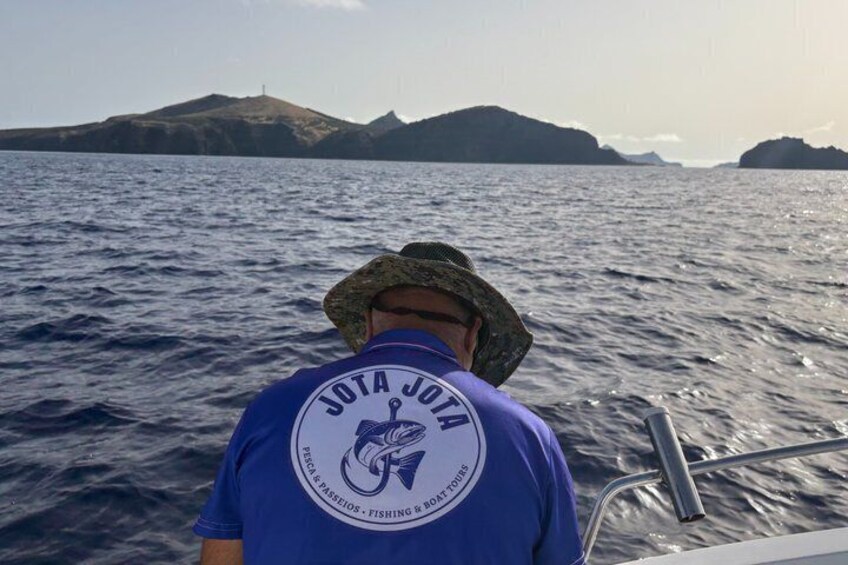 Una persona con una camisa azul que lleva un logotipo y texto en la parte trasera, de pie en un bote con una masa de agua y accidentes geográficos distantes al fondo. 1