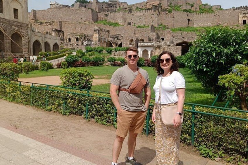 A man and a woman standing in front of a historic fort with greenery and a clear sky. 1