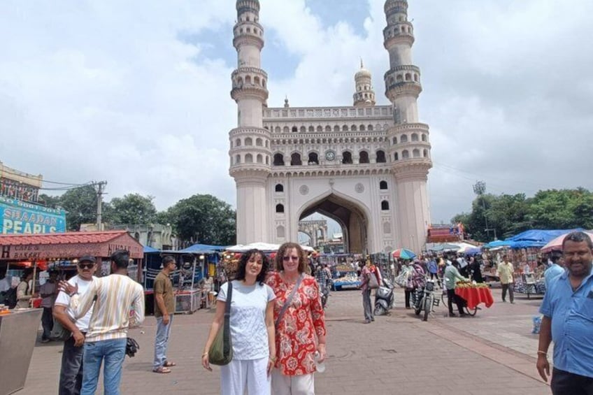 A historic building with two minarets, a bustling market with people and stalls, and a clear sky. 4