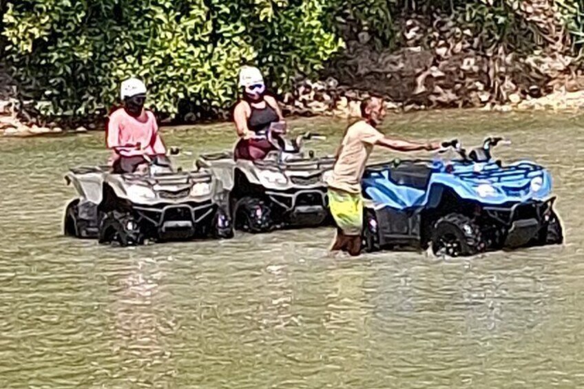Three people on ATVs in a body of water, with a rocky shoreline in the background. 1