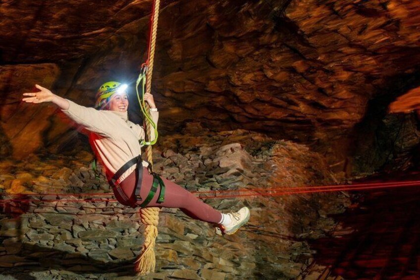 A person suspended in mid-air, wearing a harness and helmet, in a rocky cavern. 4